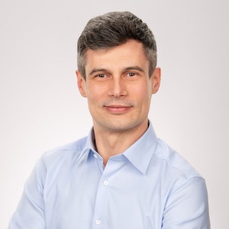 Mann mit kurzem, grauem Haar und blauem Hemd, lächelnd vor hellem Hintergrund.